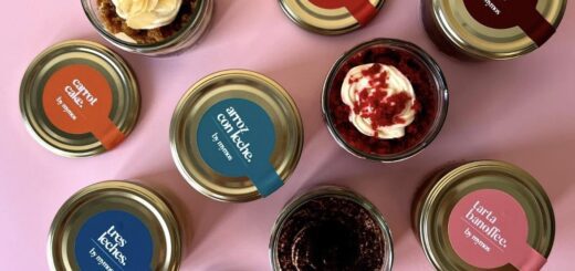 Assorted dessert jars arranged on a pink surface; labels include red velvet, carrot cake, choco cake, tiramisu, and tres leches flavors visible on the lids, with whipped toppings in some jars.