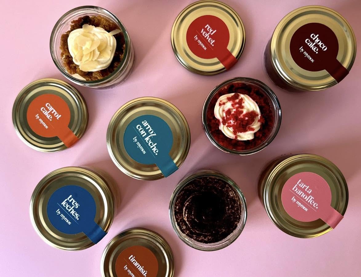 779031 - Mymos | ABC24 Noticias Assorted dessert jars arranged on a pink surface; labels include red velvet, carrot cake, choco cake, tiramisu, and tres leches flavors visible on the lids, with whipped toppings in some jars.