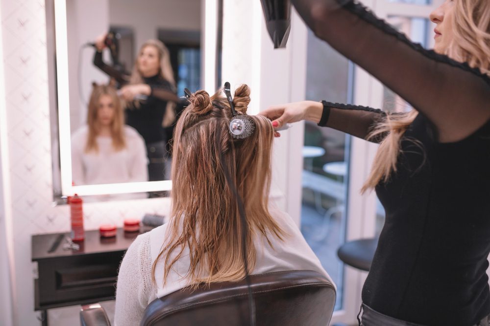 Los servicios de peluquería amplían sus propuestas para responder a nuevas necesidades - 780247 - L-equip | ABC24 Noticias Los servicios de peluquería amplían sus propuestas para responder a nuevas necesidades - Hairstylist curling a client's hair with rollers in a bright salon, reflected in the mirror behind them.