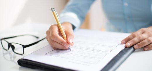 Person in a blue shirt signs a document on a clipboard while glasses lie on the white desk nearby
