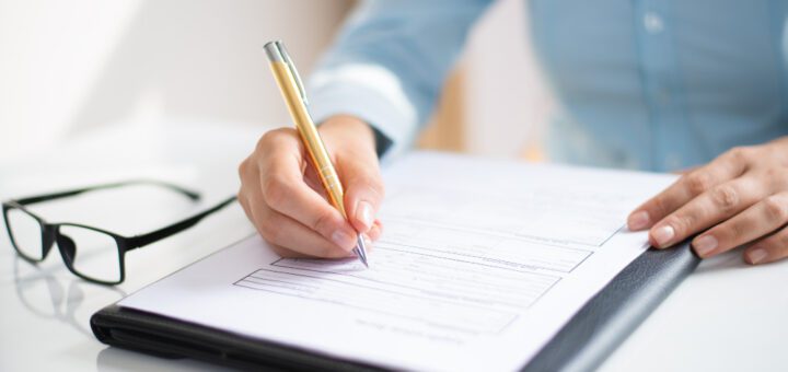 Person in a blue shirt signs a document on a clipboard while glasses lie on the white desk nearby