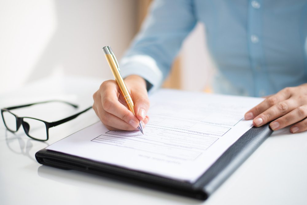 Person in a blue shirt signs a document on a clipboard while glasses lie on the white desk nearby